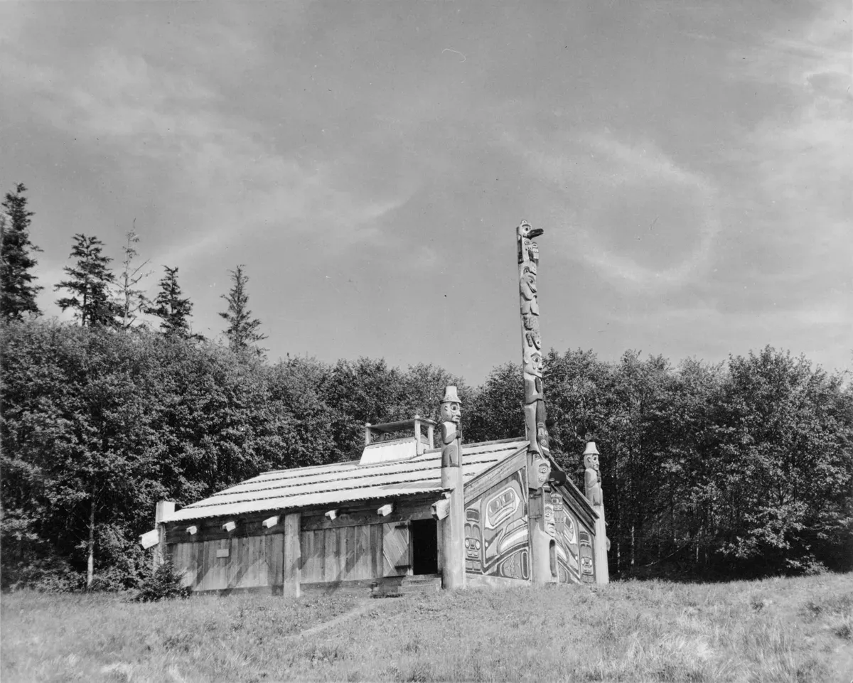 The Totem Bight community house with its painted formline front and a carved pole beside it, photographed at Totem Bight State Historical Park near Ketchikan in 1970